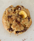 A single chocolate chip cookie with a banana chunk, photographed from directly above on a light beige surface.