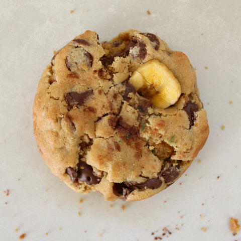 A single chocolate chip cookie with a banana chunk, photographed from directly above on a light beige surface.